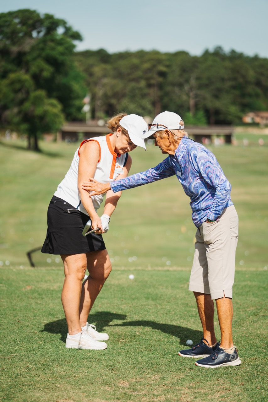 A golf instructor demonstrating a swing technique to a student on a sunny day. The student, wearing a white top and black skirt, receives guidance as they stand on a grassy range, surrounded by trees.