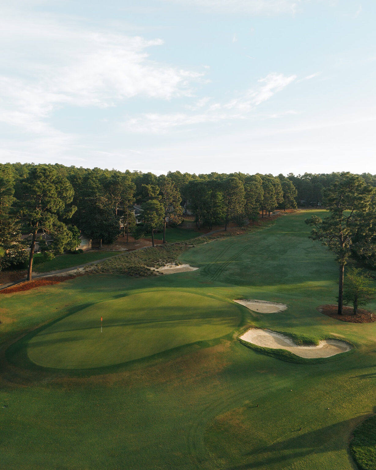 An aerial view of a golf course featuring a green with a flagstick, sand bunkers to the right, and a tree-lined background under a blue sky with scattered clouds. The manicured grass is in shades of green, indicating a well-maintained landscape.