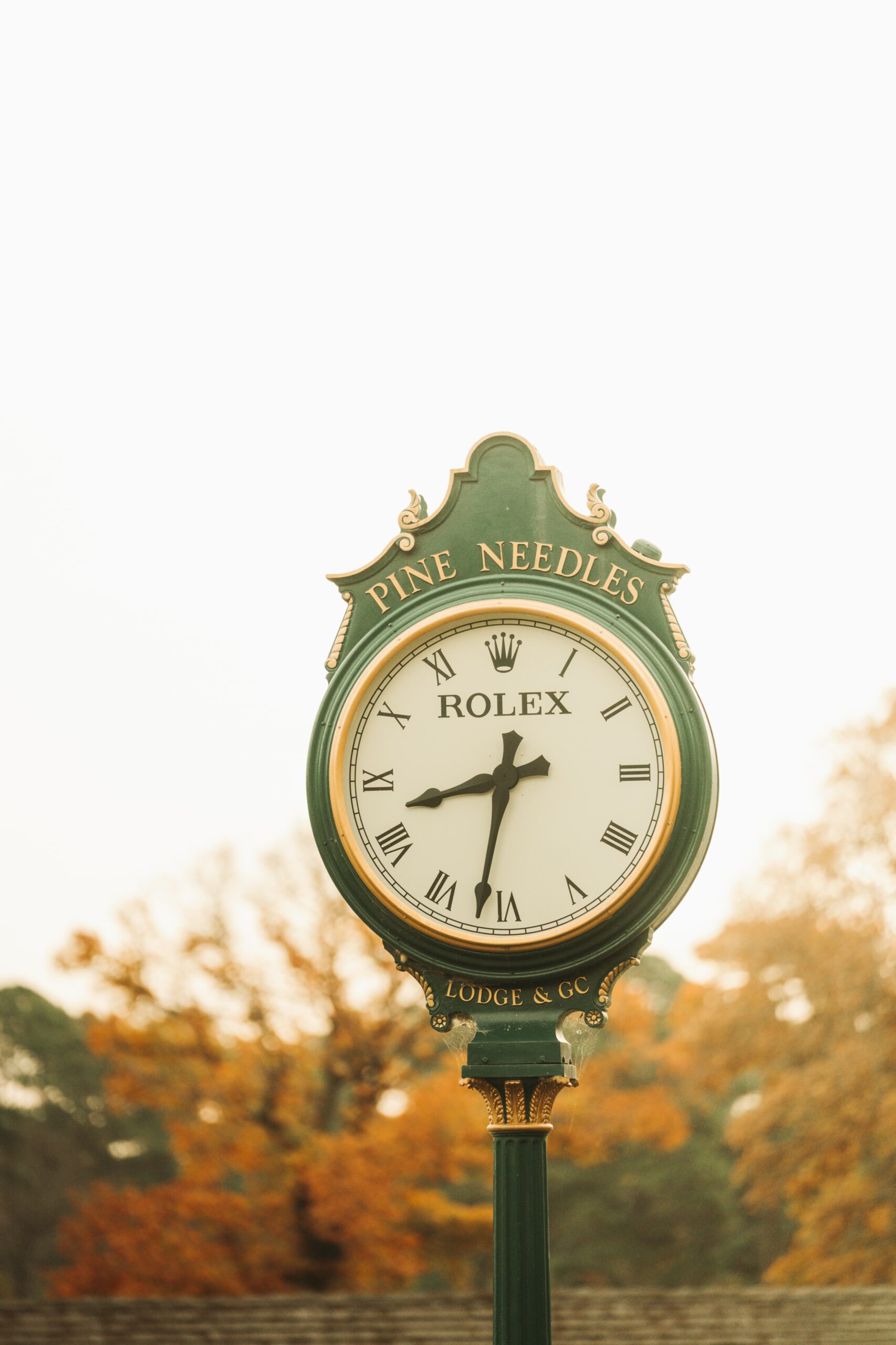 A decorative green clock with a white face displaying Roman numerals. The clock is marked with the brand name "Rolex" and the text "Pine Needles Lodge & Co." in gold lettering at the top. Autumn foliage is blurred in the background.