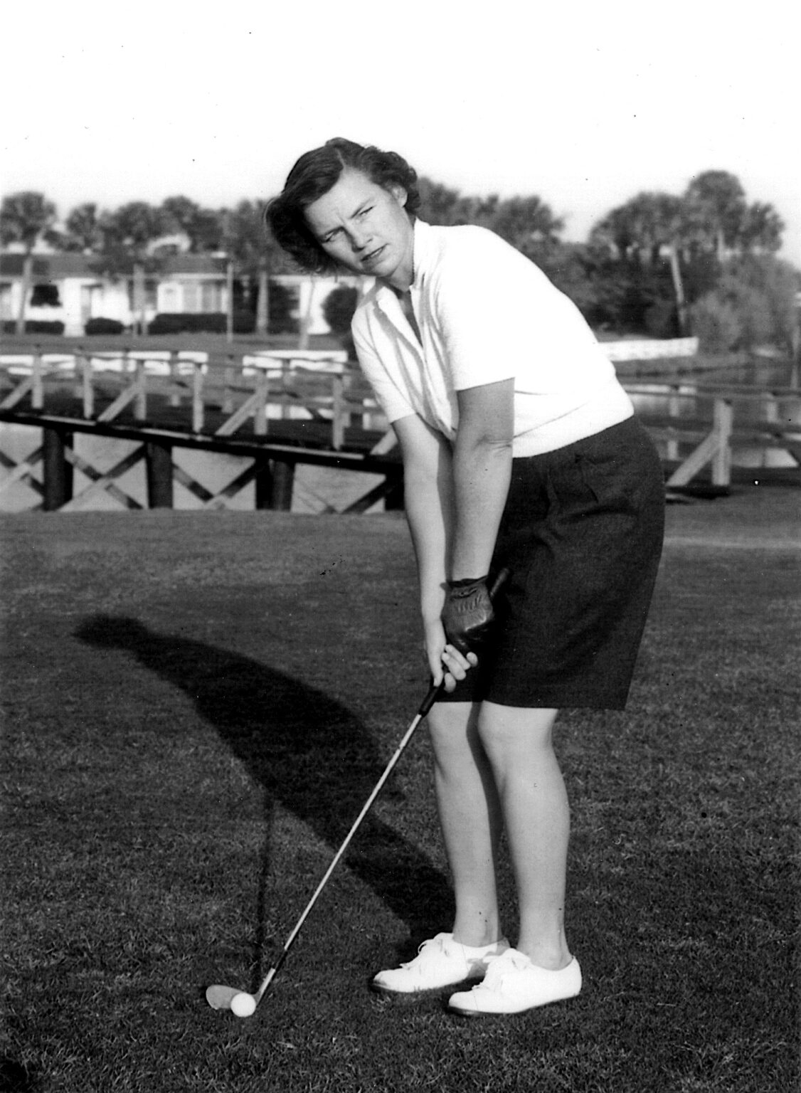 A young woman dressed in a white blouse and dark skirt stands on a golf course, preparing to putt. She has short hair and is focused on her stance, with a putter ready near a small golf ball. In the background, a wooden bridge and trees are visible.