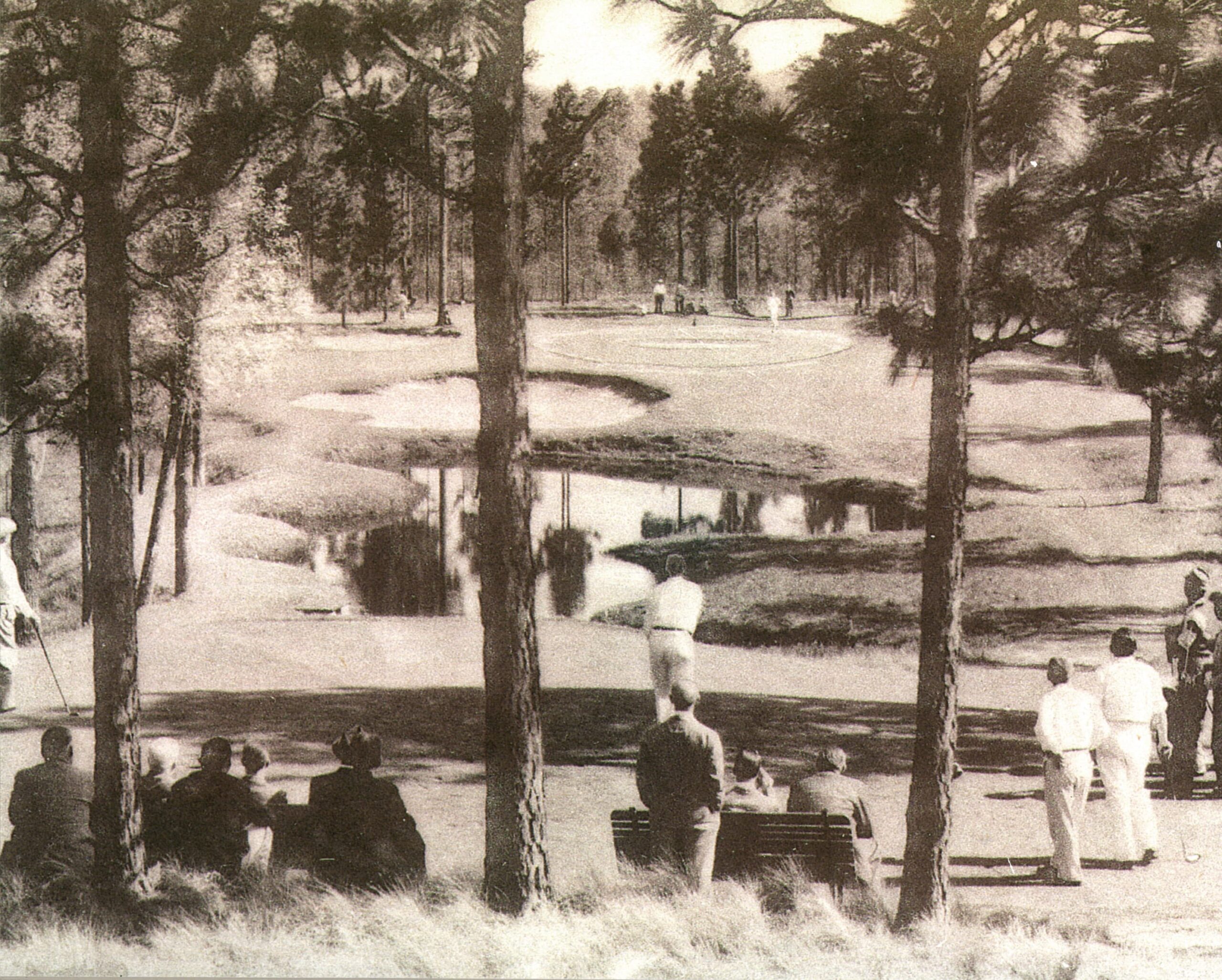 A black and white photograph showing a golf course with several players on the green. Spectators are seated on benches under tall trees near a water hazard. The scene captures a peaceful day on the course, with a view of the landscape and players.