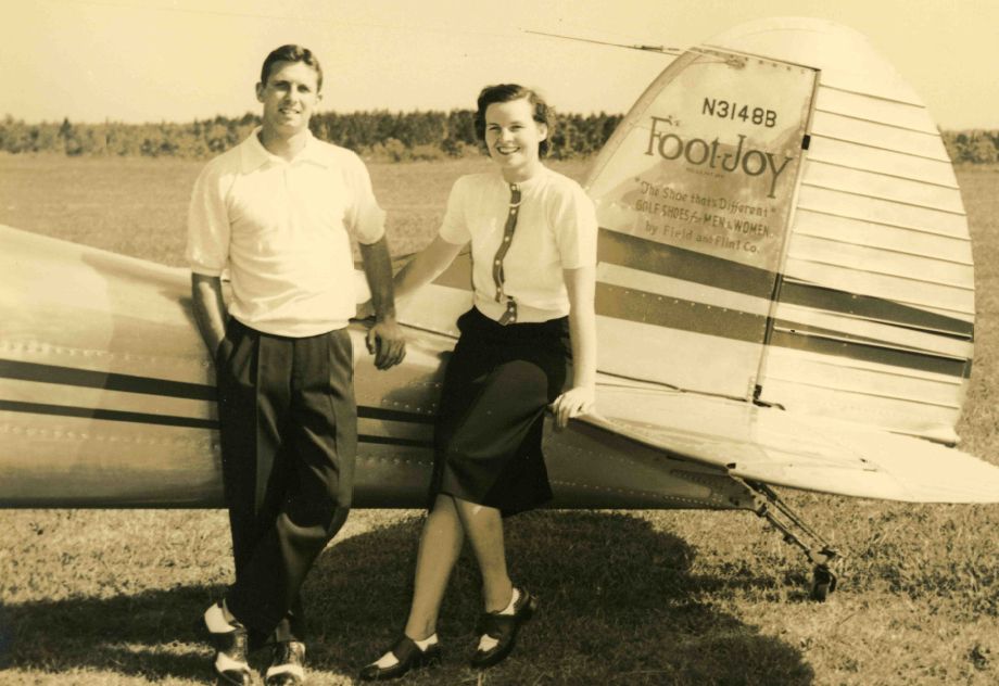 A man and a woman stand beside a vintage airplane on a grassy field. The man wears a white shirt and dark pants, while the woman wears a white blouse and a dark skirt. Both are smiling at the camera. The airplane displays the name 'FootJoy' and has a striped design.