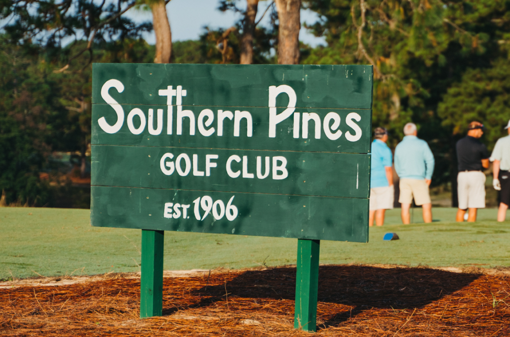 A wooden sign for Southern Pines Golf Club displayed on the golf course. The sign features white text on a green background, stating the club's name and 'EST. 1906.' In the background, several people are seen preparing to play golf.