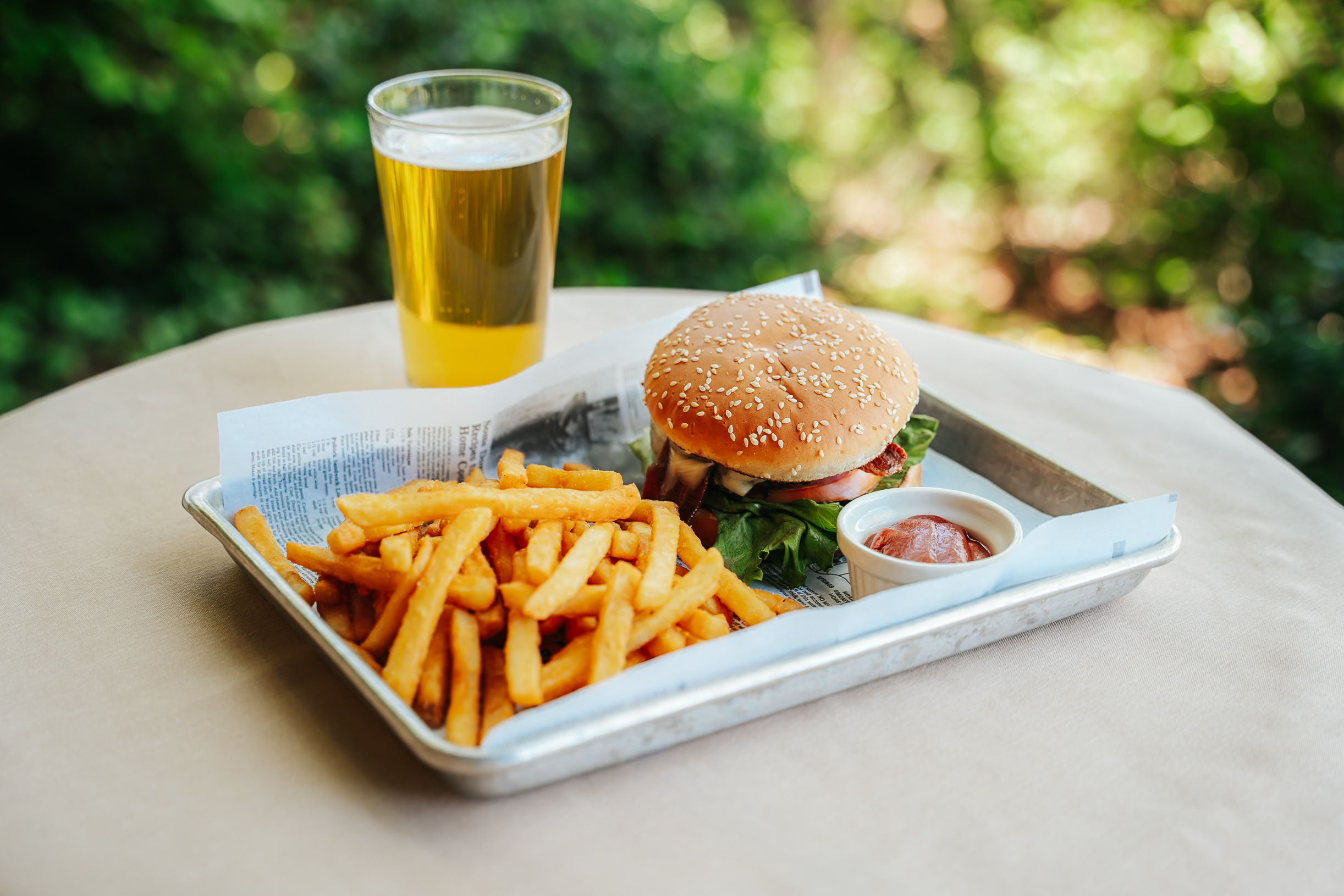 A tray containing a sesame seed bun burger with lettuce, tomato, and onion, served with golden French fries and a small dish of ketchup. A pint of beer is placed next to the tray, all set on a table with a natural background.