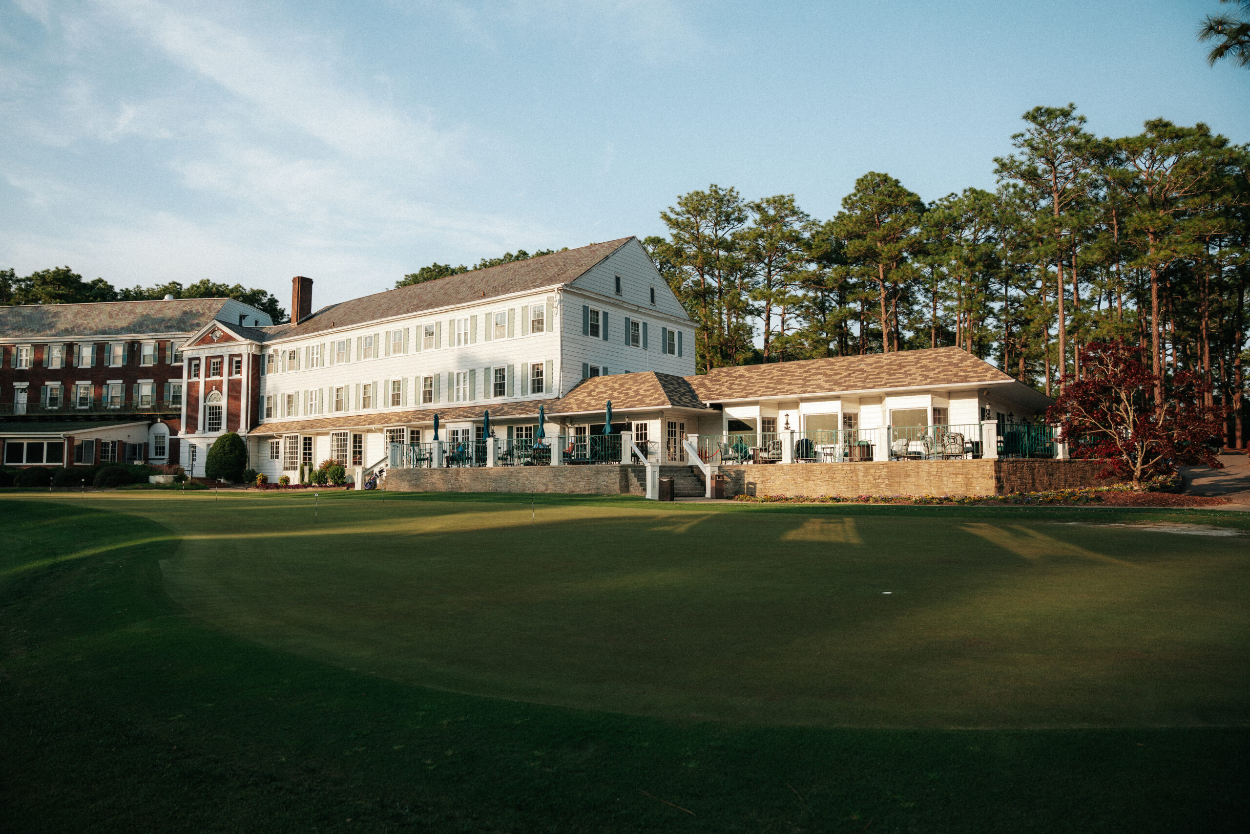 A large two-story building with multiple windows and a sloped roof is set near a golf course. The structure is surrounded by trees, and a patio area is visible. Sunlight casts shadows on the well-maintained grass in the foreground.