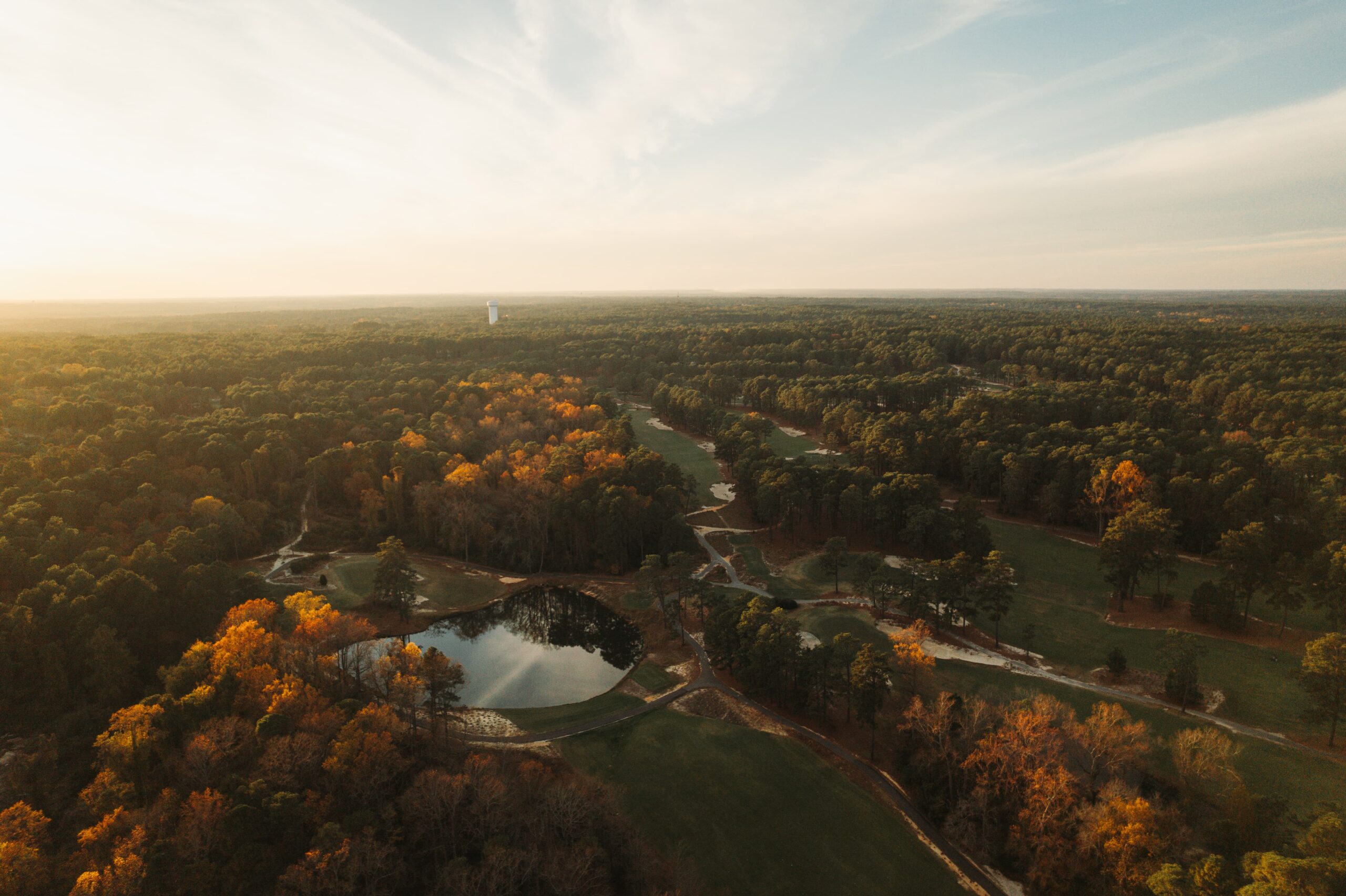 An aerial view of a landscape featuring a mixture of green trees and autumn foliage. A winding creek flows through the scene, with a small pond reflecting the surroundings. The sky is light with soft clouds, and the image captures a tranquil natural setting.