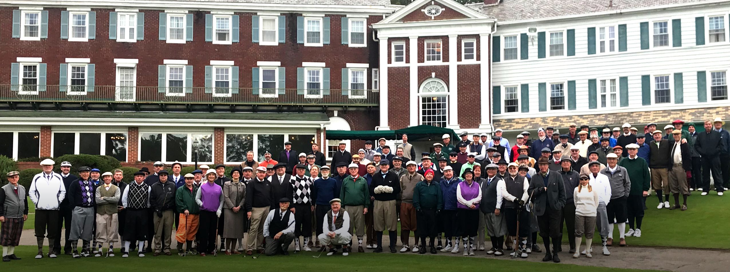 A large group of men and women gathered on a golf course in front of a historic clubhouse. The individuals are dressed in typical golf attire, including caps and sweaters, and are positioned on a green lawn, showcasing the architecture of the building behind them.