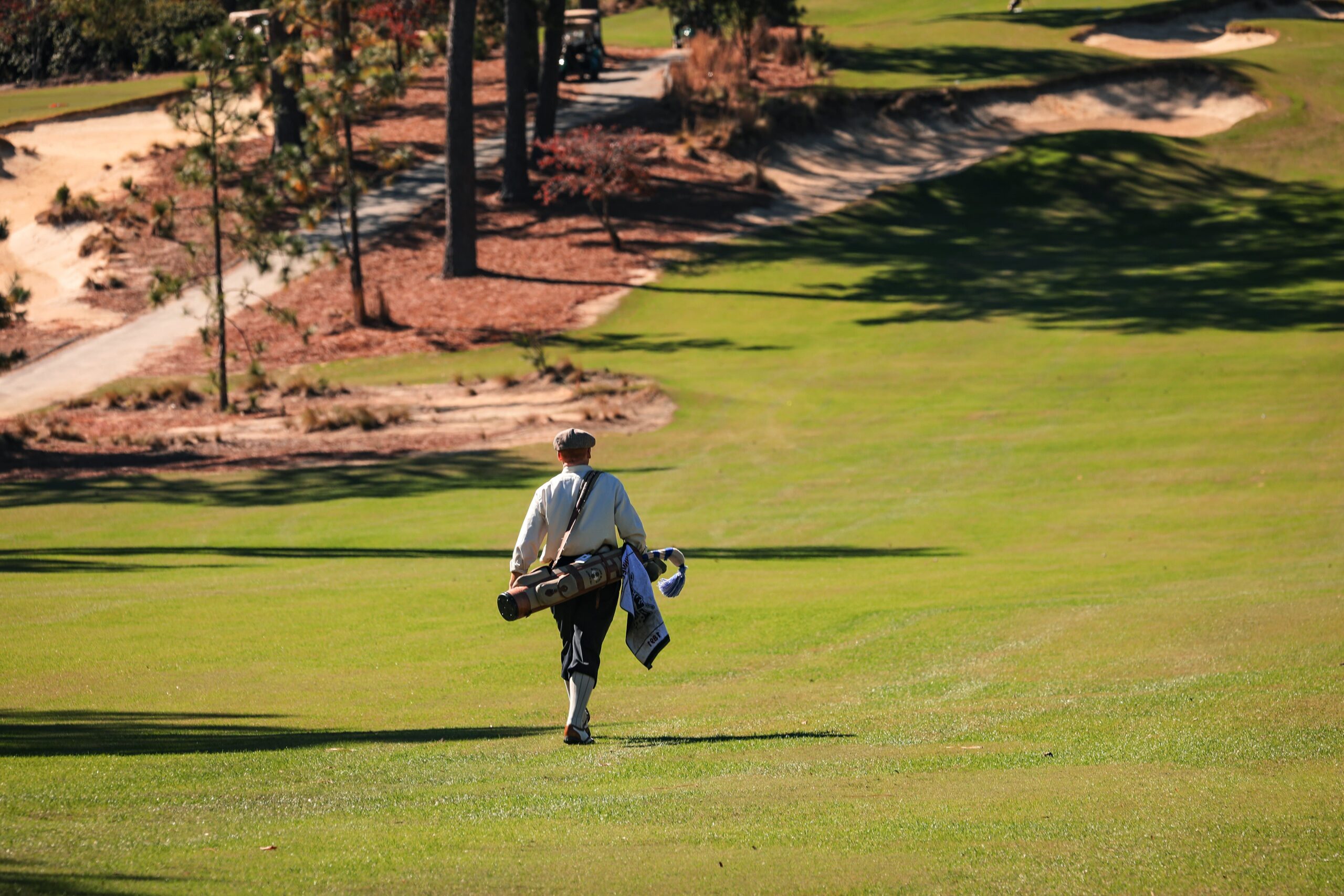 A golfer walking on a lush green golf course, carrying a golf bag over their shoulder. The background features a winding path and trees, indicating a well-maintained outdoor setting.