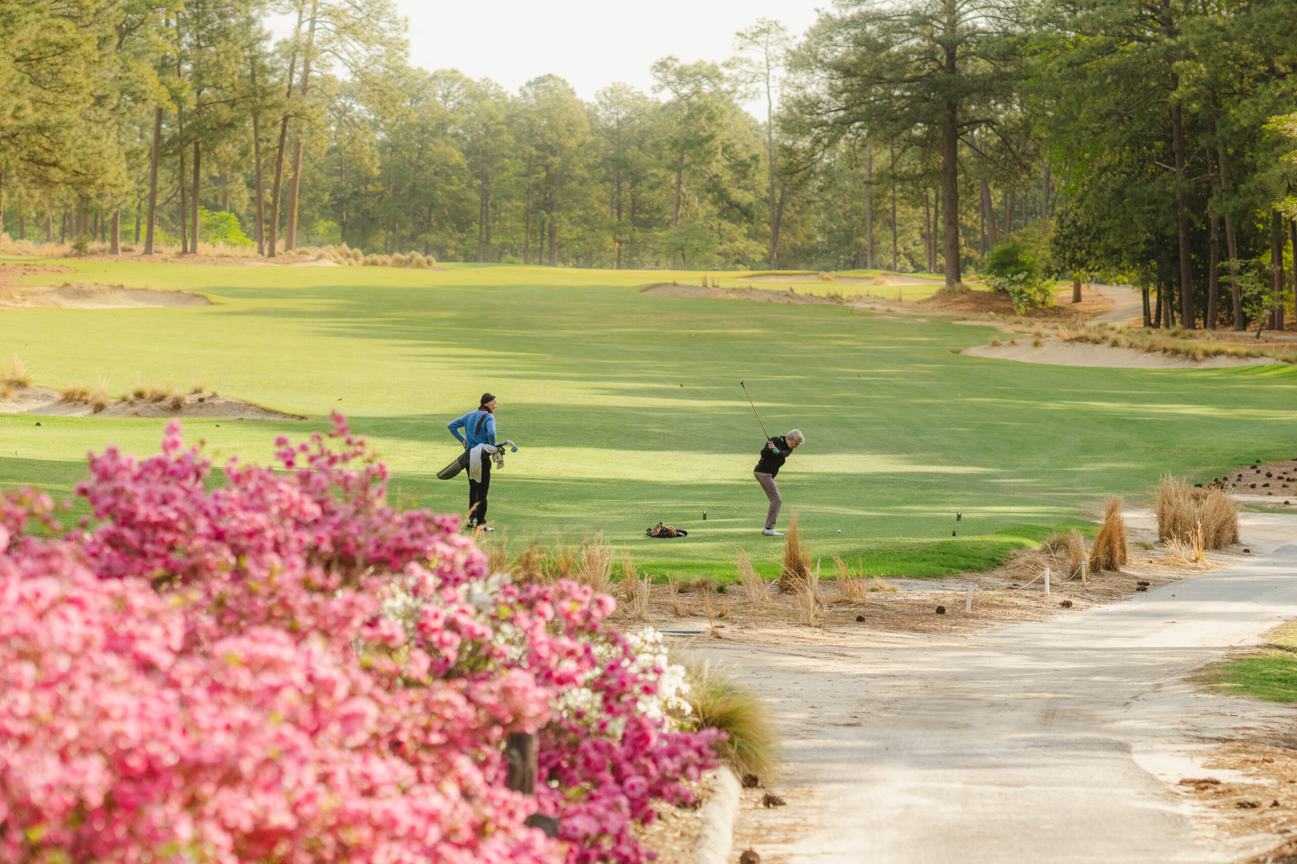 Two golfers playing on a lush, green fairway, surrounded by tall trees. In the foreground, vibrant pink flowers add color to the scene. The atmosphere is calm, with wide open spaces visible in the background.