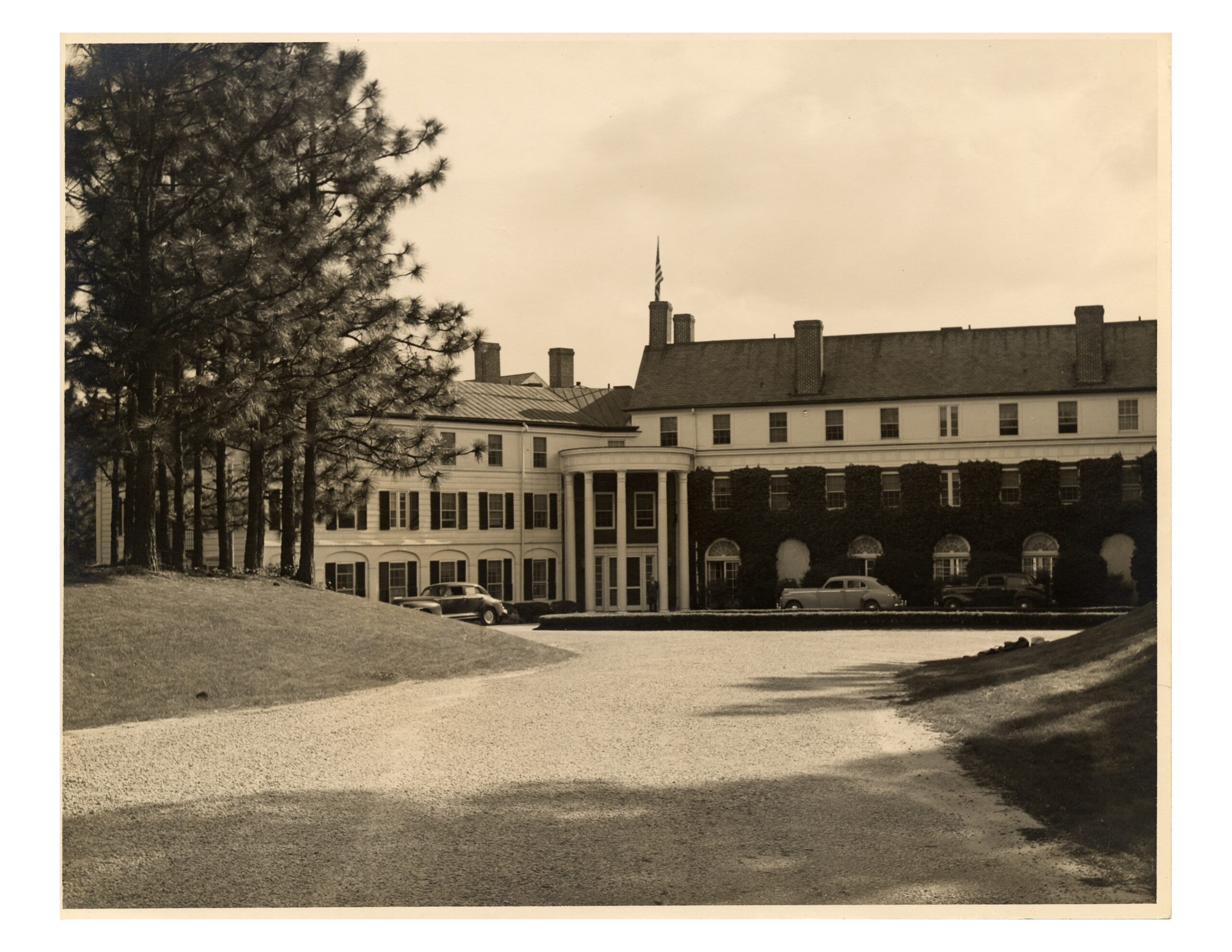 A historic building with a prominent white entrance and columns, surrounded by trees, seen from a gravel driveway. The structure features multiple windows and a sloped roof, reflecting early 20th-century architectural style.