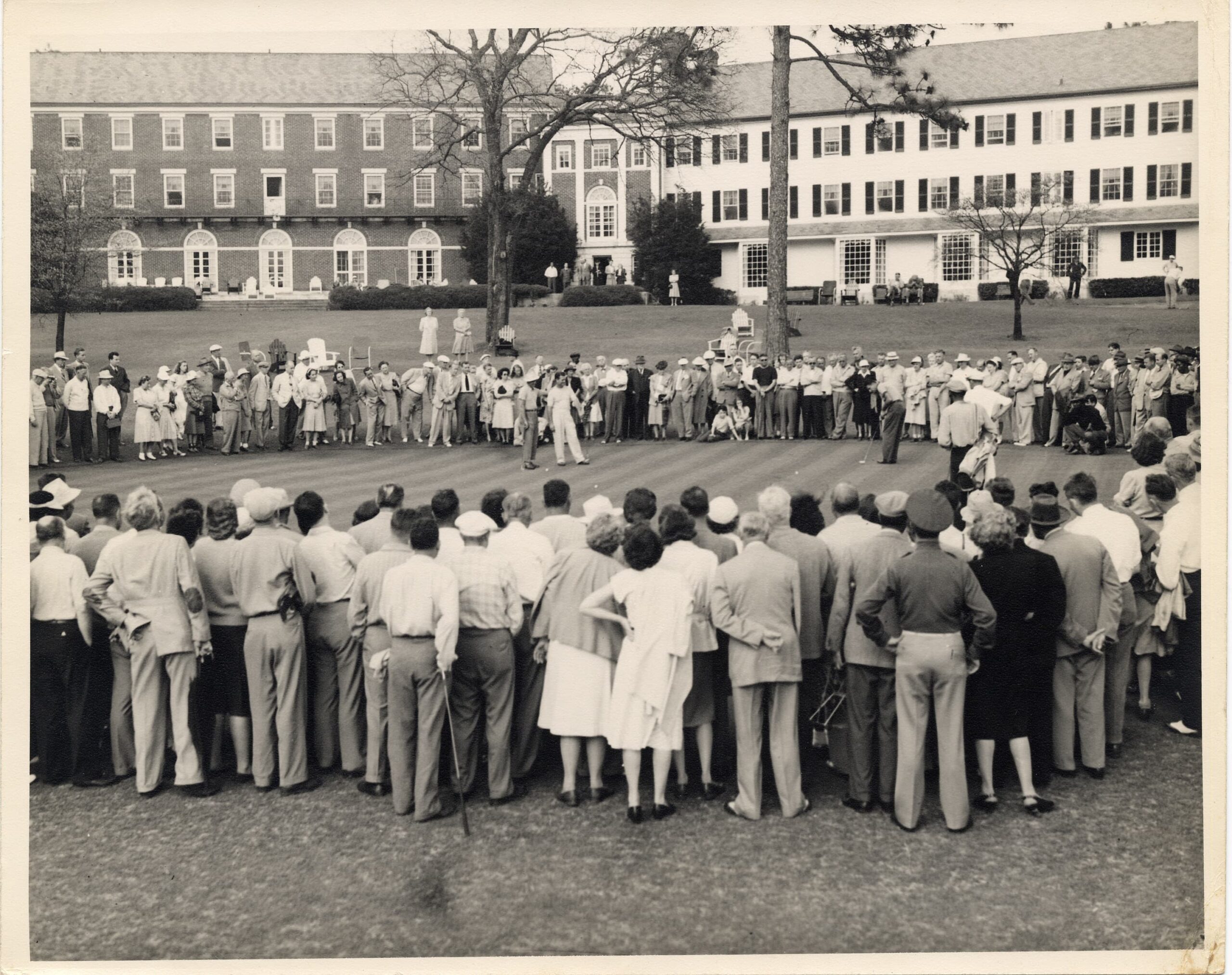 A large crowd of people gathered on a grassy area, observing an event taking place on a putting green. The background features two multi-story buildings, with people in period clothing from the mid-20th century standing in close formation.