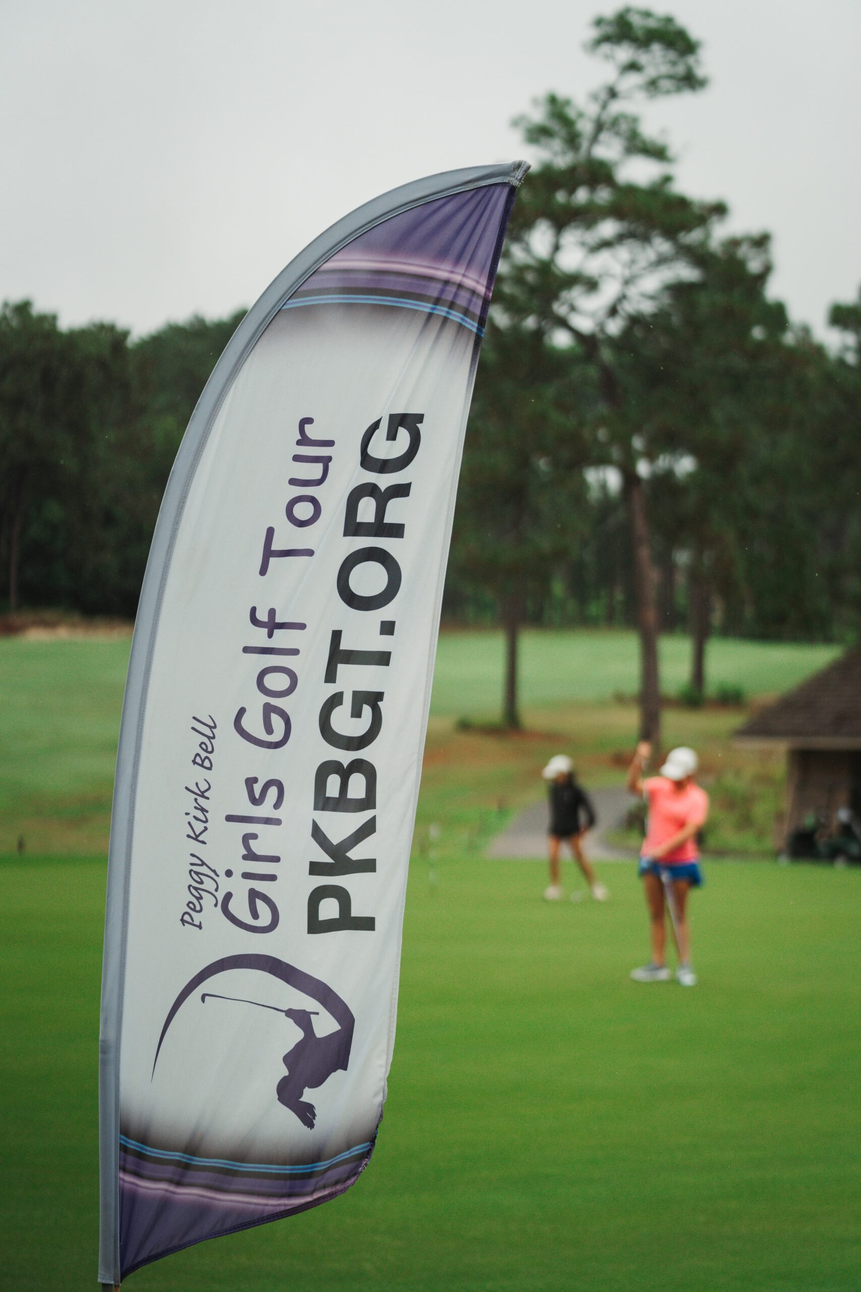 A banner for the Peggy Kirk Bell Girls Golf Tour stands prominently on the golf course, displaying the website PKBGT.ORG. In the background, female golfers are seen practicing on the green, surrounded by trees.