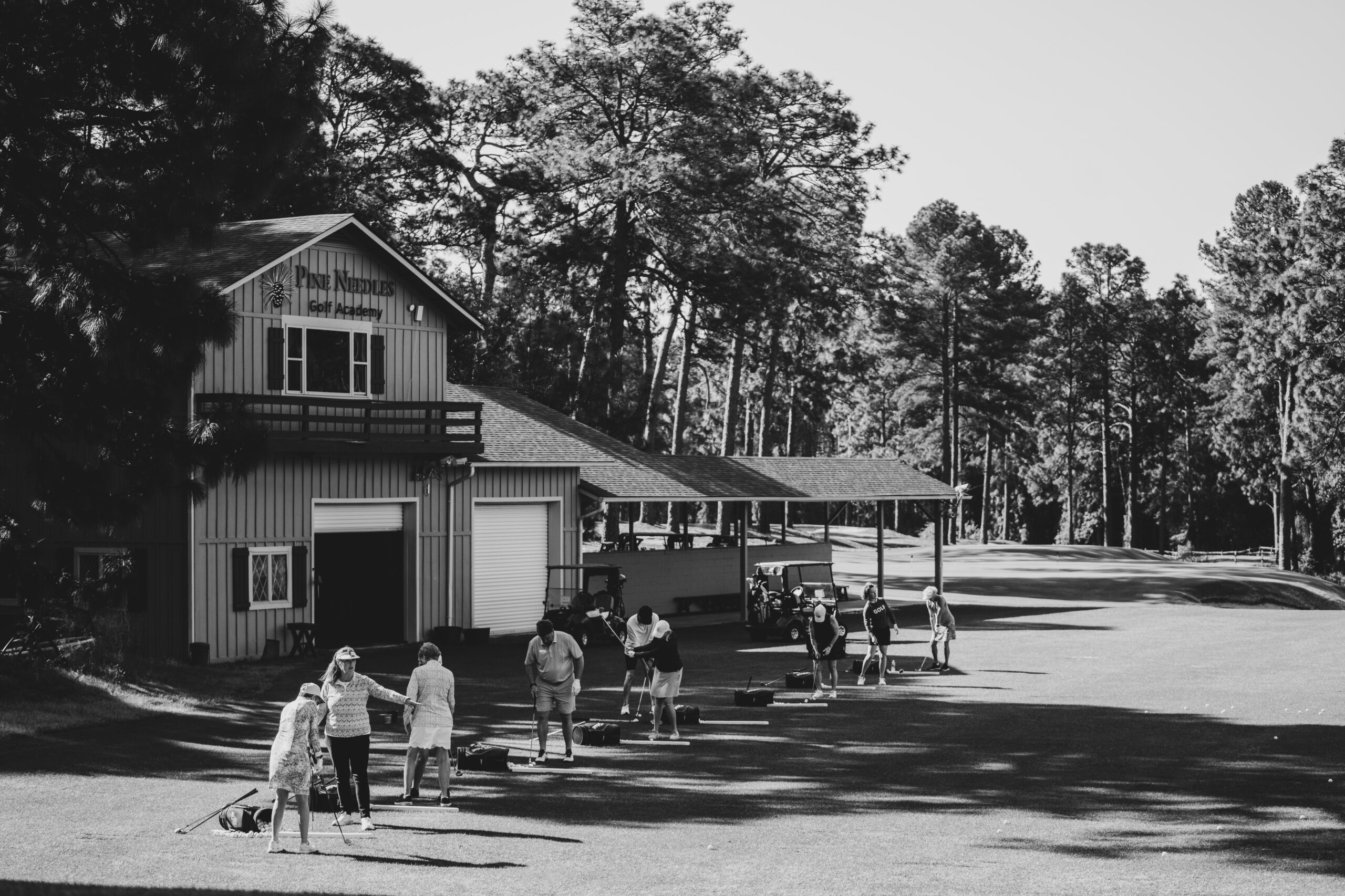 A black-and-white photo of a golf practice area featuring a wooden building with a veranda. Several people are engaged in activities on the grass in front of the building, surrounded by tall trees.