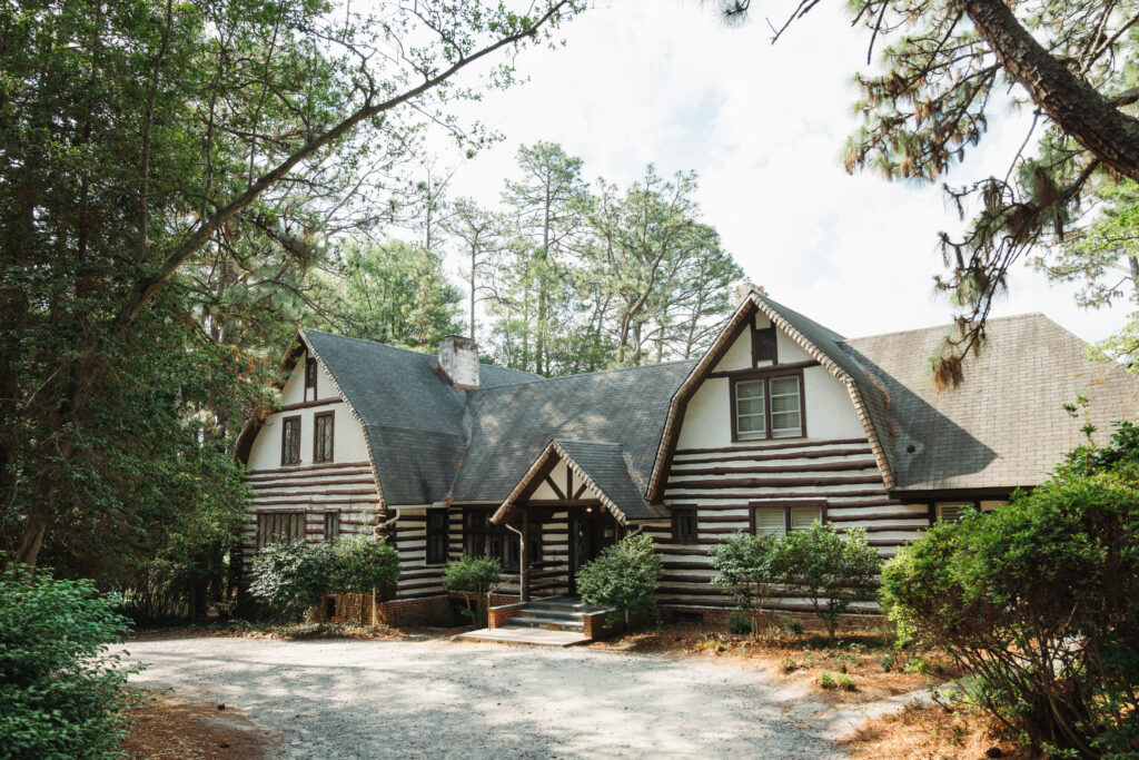 A rustic log cabin with steep gabled roofs sits among tall pine trees, with a gravel driveway and shrubs in the foreground.